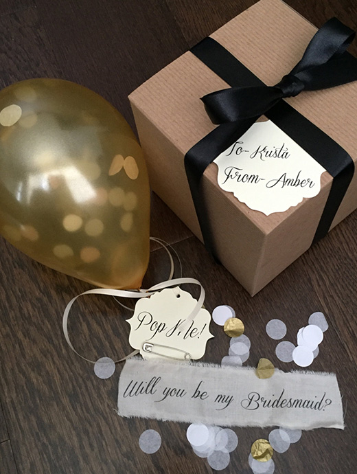 A confetti box filled with a balloon and a bridesmaid proposal written on ribbon