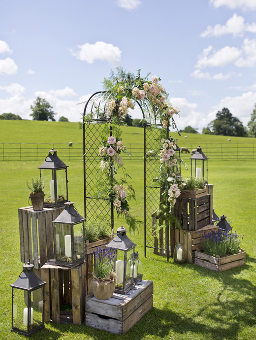 A metal wedding arch with floral décor surrounded by boxes, plants and lanterns